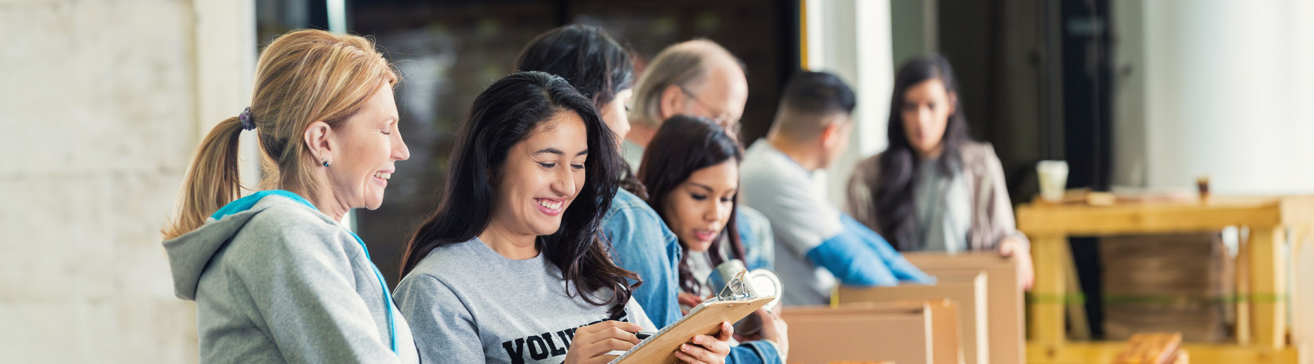 Mature adult Caucasian woman is standing with mid adult Hispanic woman as they pack cardboard boxees full of donated food in charity food bank. Other volunteers are lined up behind them, also sorting donated groceries into boxes. Hispanic woman is writing on checklist on clipboard.