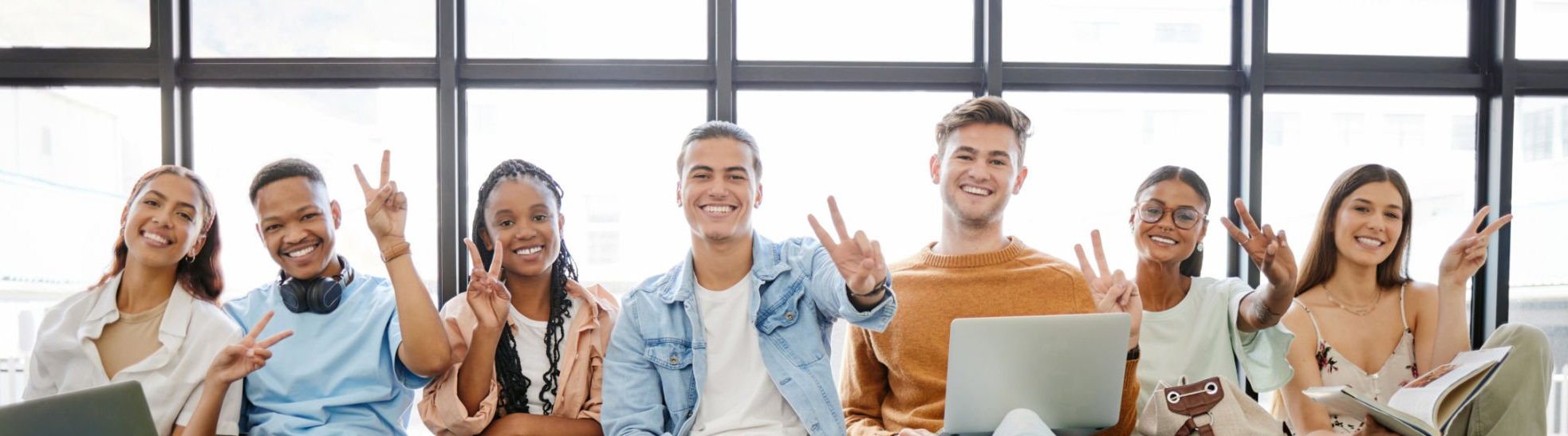 A group of teens/young adults sitting with their backs against a window and laptops on their laps, making the peace sign