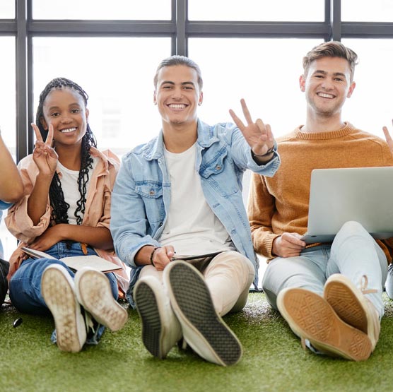 A group of teens/young adults sitting with their backs against a window and laptops on their laps, making the peace sign