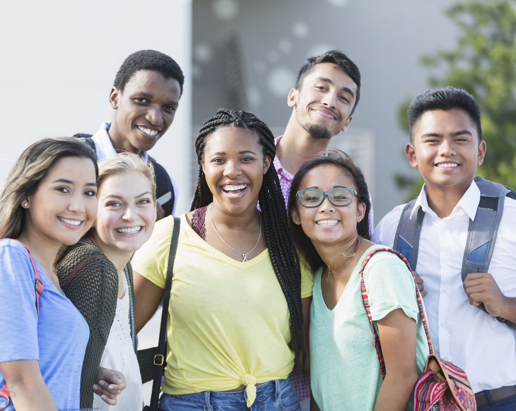 A group of eight multi-ethnic teenagers, 17 and 18 years old, carrying book bags, standing together outside a school building. They are high school seniors or university freshmen.